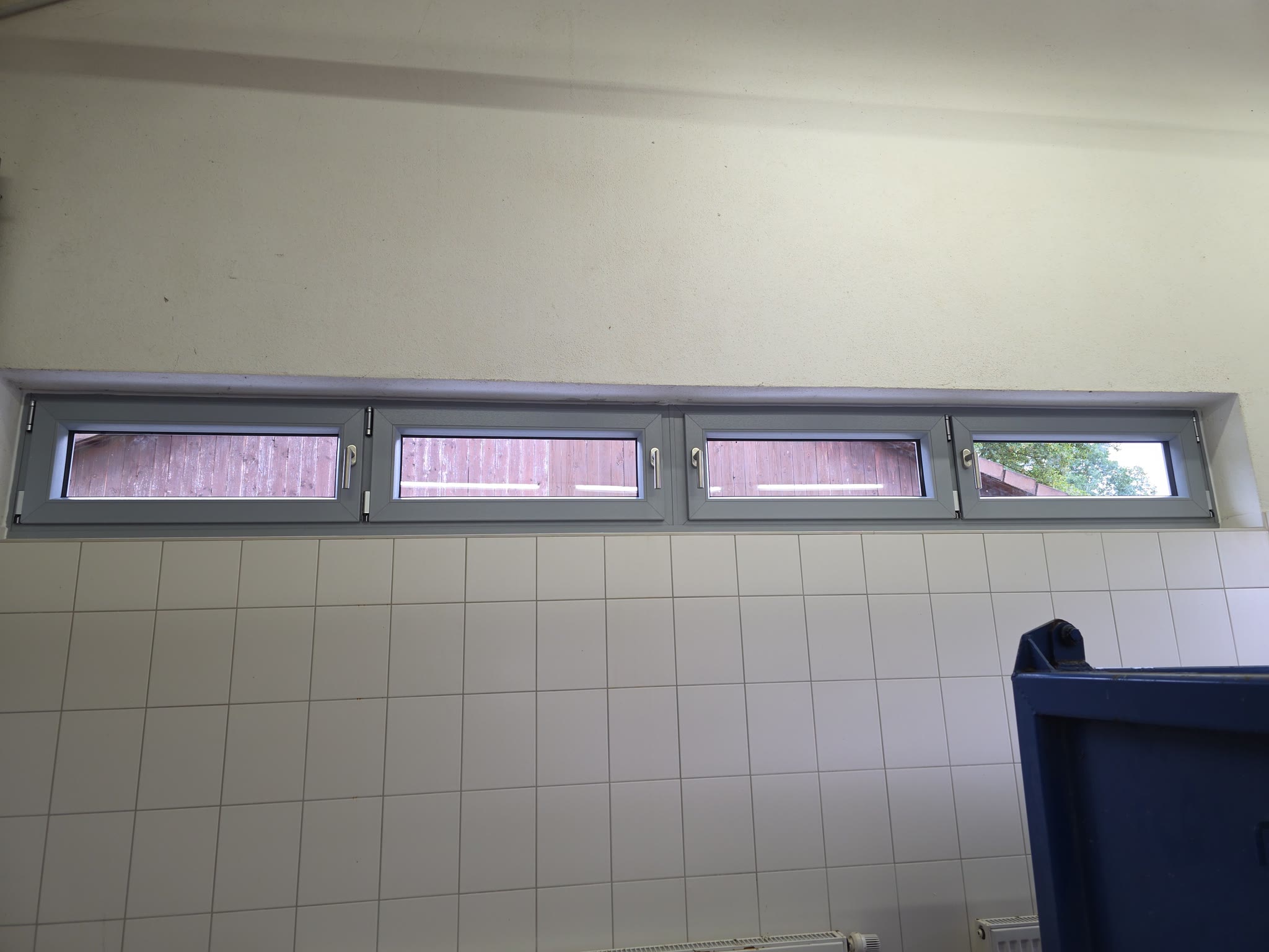 Vierteiliges Fensterband mit montierten Beschlägen und fertig gestrichener Laibung, Blick auf die Holzfassade gegenüber.