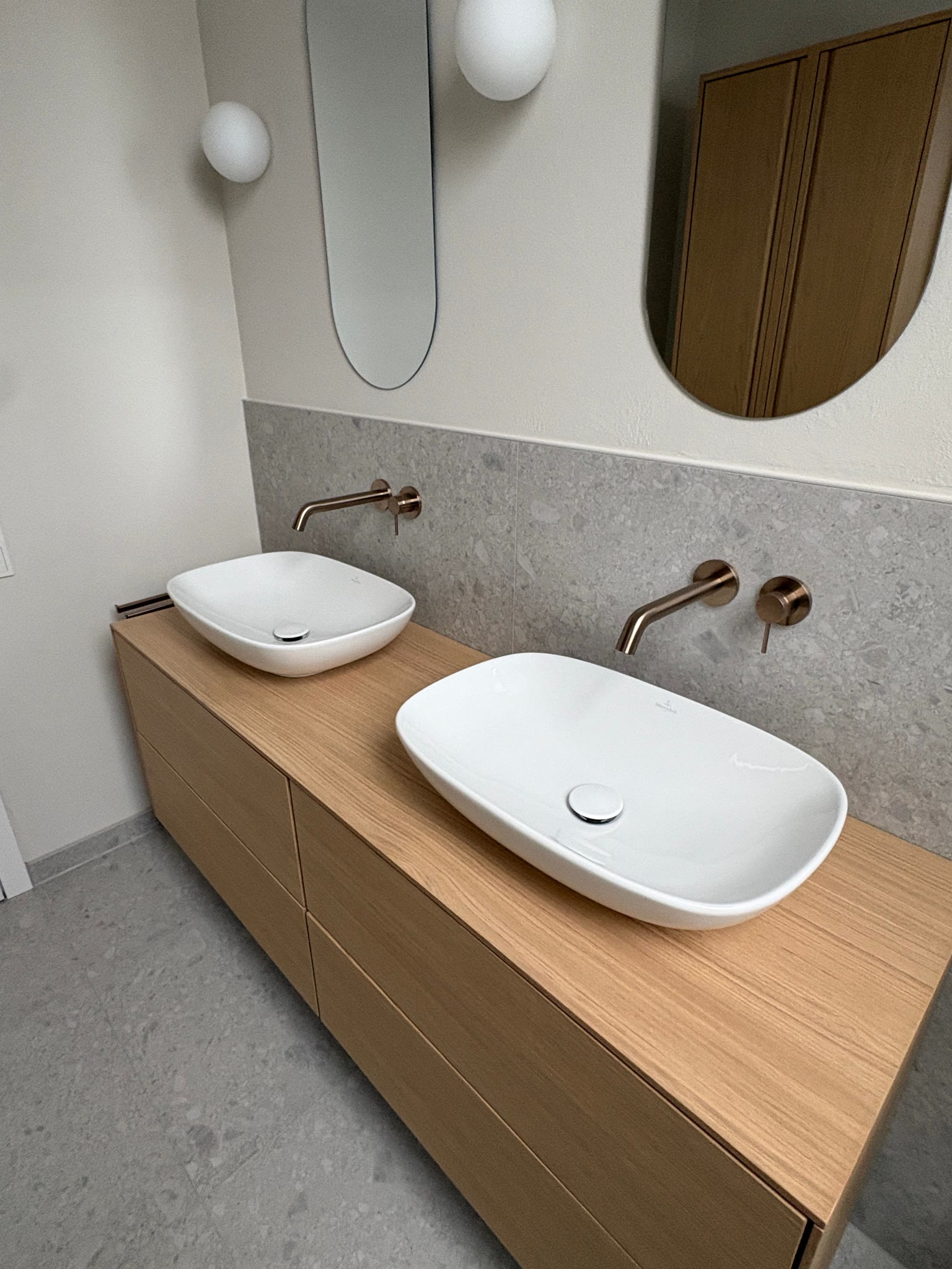 Freitragender Eichen-Waschtisch mit zwei Aufsatzbecken und bronzefarbenen Wandarmaturen vor grauer Steinoptik.